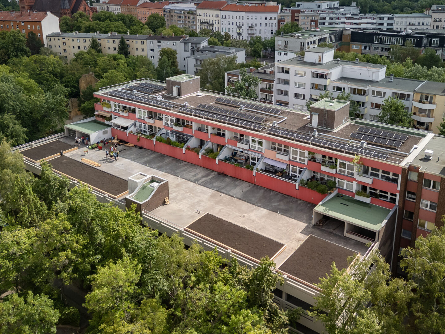 Luftaufnahme eines Wohnblocks mit Solaranlagen auf dem Dach; auf dem Vordach bereiten Menschen lange Beete für einen Gemeinschaftsgarten vor.