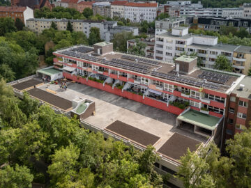 Luftaufnahme eines Wohnblocks mit Solaranlagen auf dem Dach; auf dem Vordach bereiten Menschen lange Beete für einen Gemeinschaftsgarten vor.