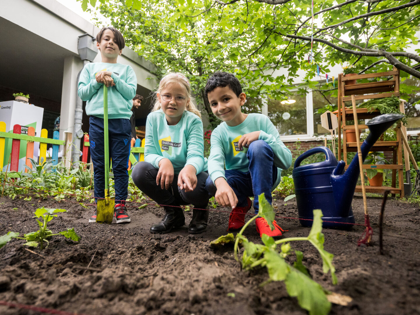 Drei Kinder in grünen Pullis gärtnern im Hof, knien am Beet mit jungen Pflanzen; Gießkanne und Werkzeug stehen bereit