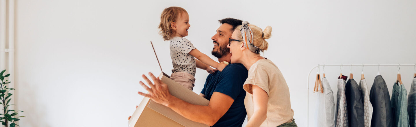Lachende Familie mit Kind hält Umzugskarton in der neuen Wohnung; Kleiderstange und Pflanze im Hintergrund, fröhliche Einzugsstimmung.