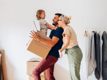 Lachende Familie mit Kind hält Umzugskarton in der neuen Wohnung; Kleiderstange und Pflanze im Hintergrund, fröhliche Einzugsstimmung.