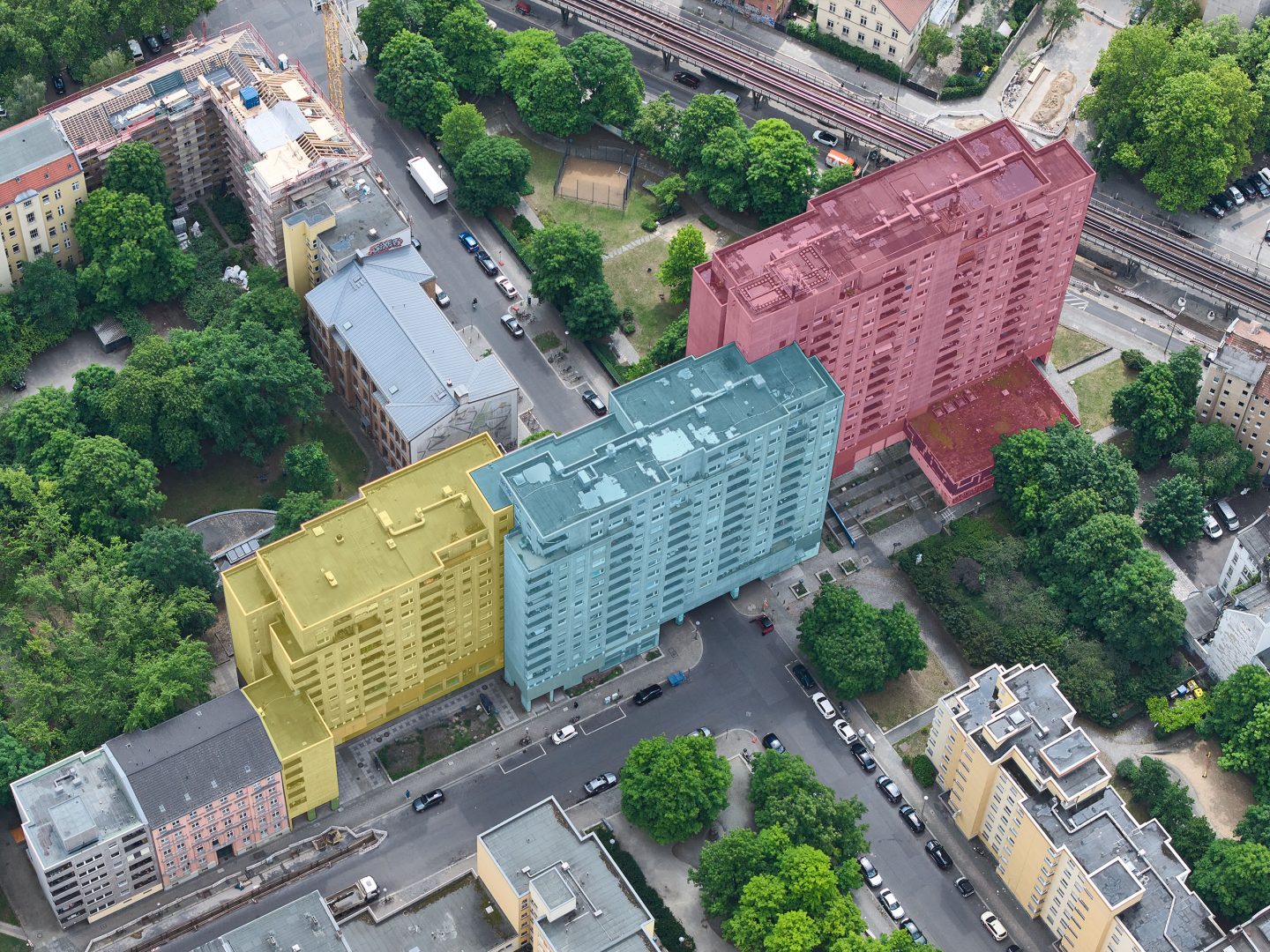 Luftaufnahme eines Stadtviertels mit drei farbig markierten Wohnblöcken: gelb, blau und rot hervorgehobene Hochhäuser zwischen Bäumen.