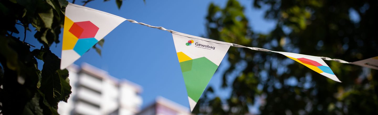 Weiße Wimpelgirlande mit buntem Gewobag-Logo hängt zwischen Bäumen im Sonnenschein, verschwommene Häuser im Hintergrund.