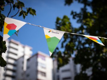 Weiße Wimpelgirlande mit buntem Gewobag-Logo hängt zwischen Bäumen im Sonnenschein, verschwommene Häuser im Hintergrund.