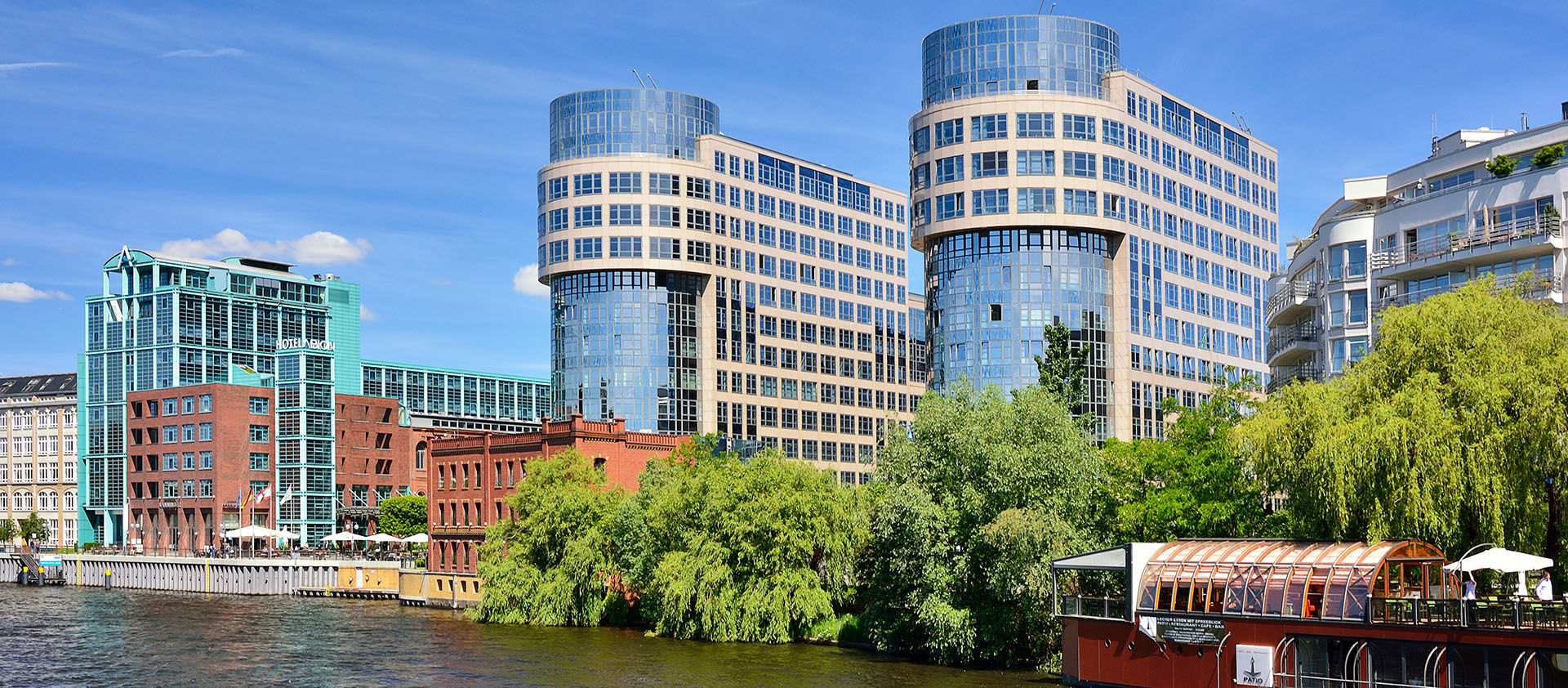 Moderne Büro- und Wohngebäude mit Glasfassaden am Flussufer, davor dichter Baumbestand und ein verglastes Schiff-Restaurant.
