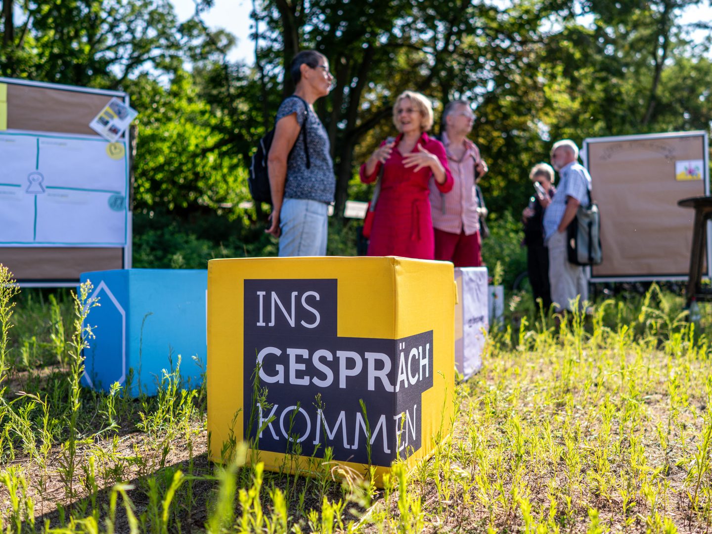 Gelber Würfel mit Aufdruck „Ins Gespräch kommen“ auf Wiese, im Hintergrund mehrere Menschen im Austausch vor Pinnwänden im Freien.