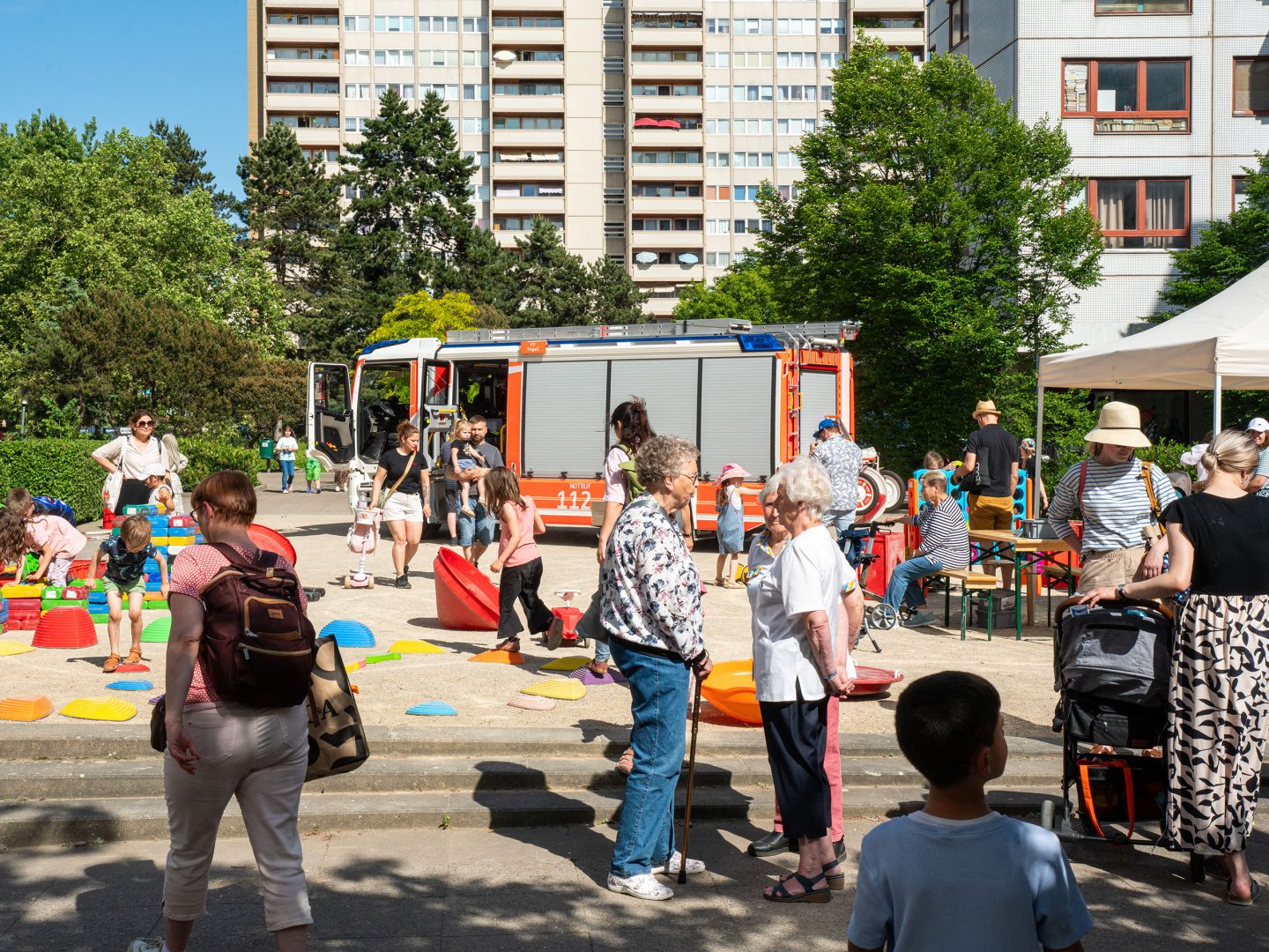 Straßenfest mit Kindern und Erwachsenen, bunter Spielbereich, Feuerwehrwagen als Attraktion, Hochhäuser und Bäume im Hintergrund.