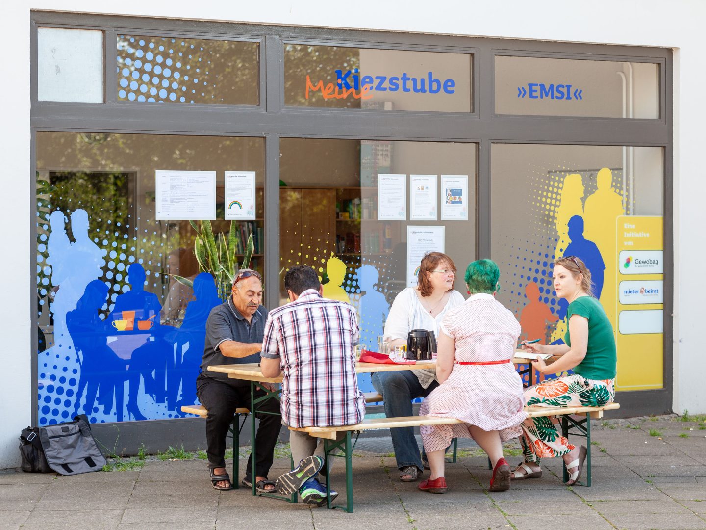 Mehrere Menschen sitzen an einem Tisch vor der bunt gestalteten „Kiezstube EMSI“ und unterhalten sich bei Getränken.