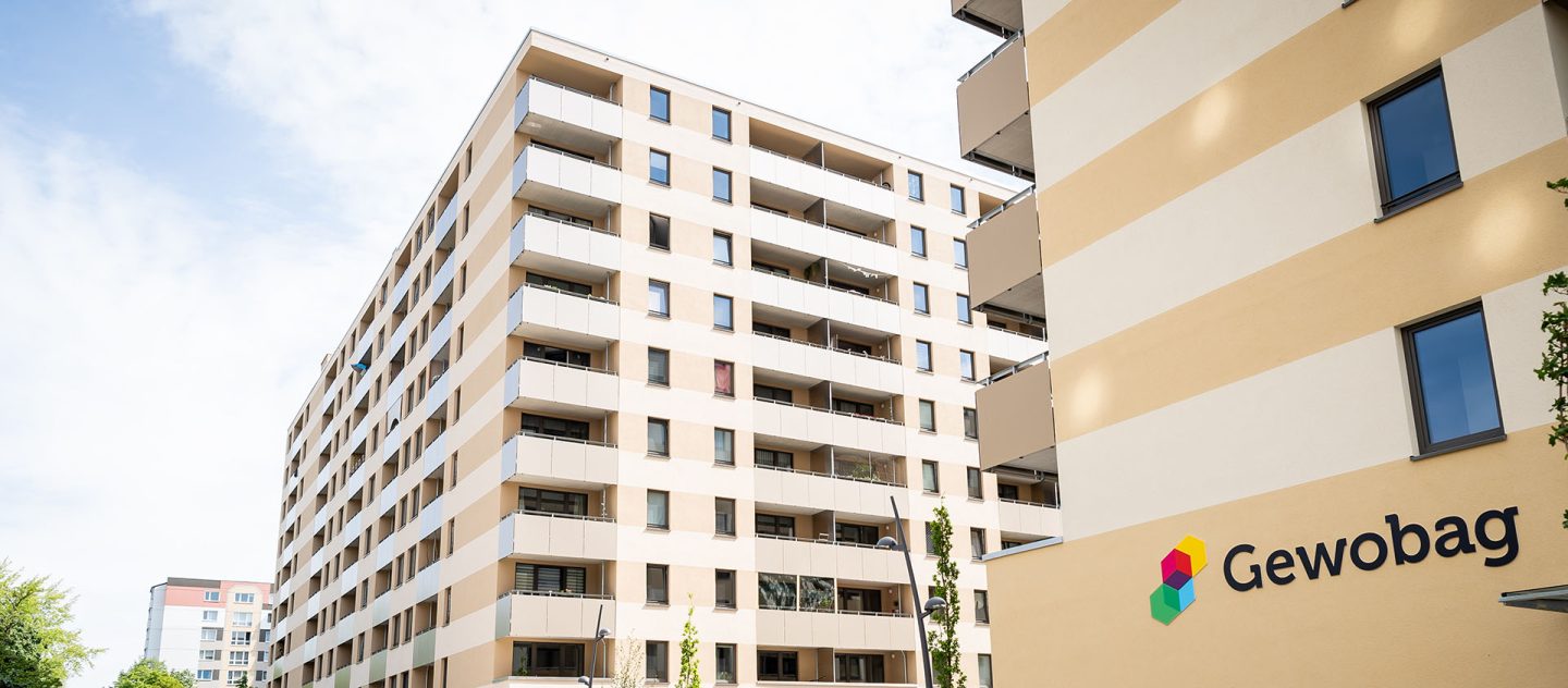 Mehrgeschossige, helle Wohnhäuser mit vielen Balkonen, rechts eine beige Fassade mit Gewobag-Logo unter leicht bewölktem Himmel.