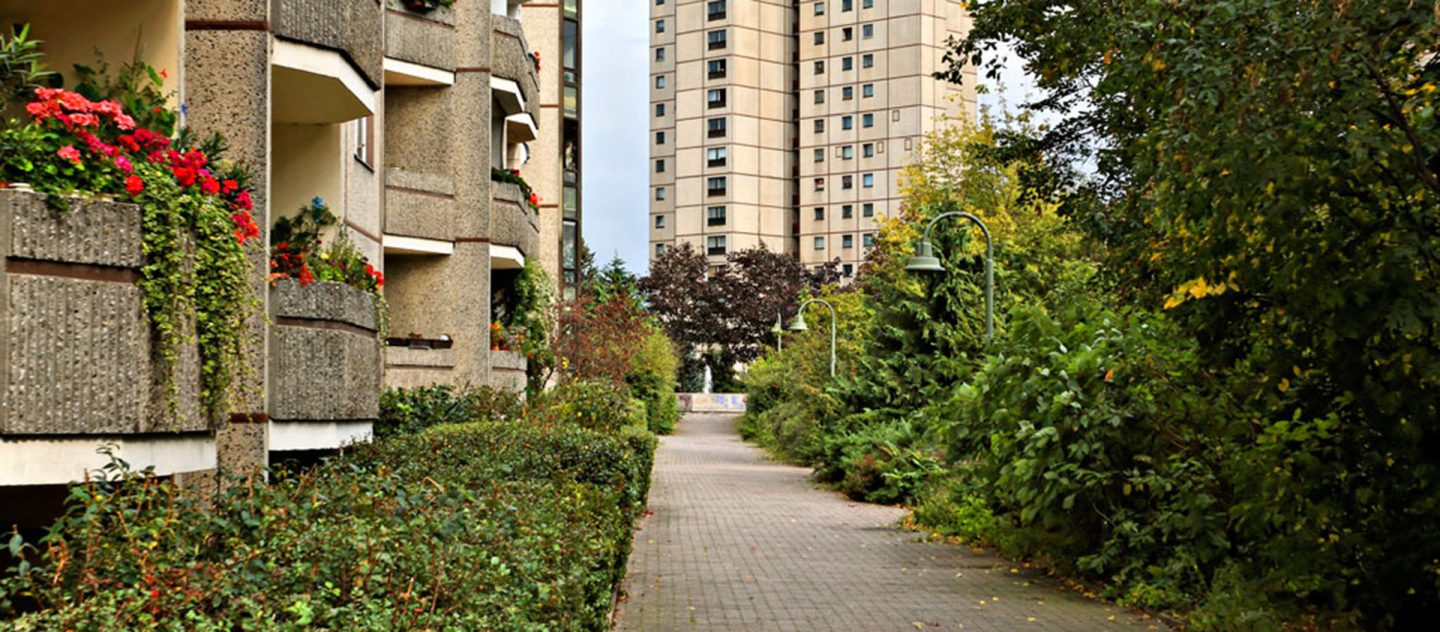 Gepflasterter Weg zwischen bepflanzten Balkonen links und dichtem Grün rechts, am Ende ein hoher Wohnblock im Hintergrund.