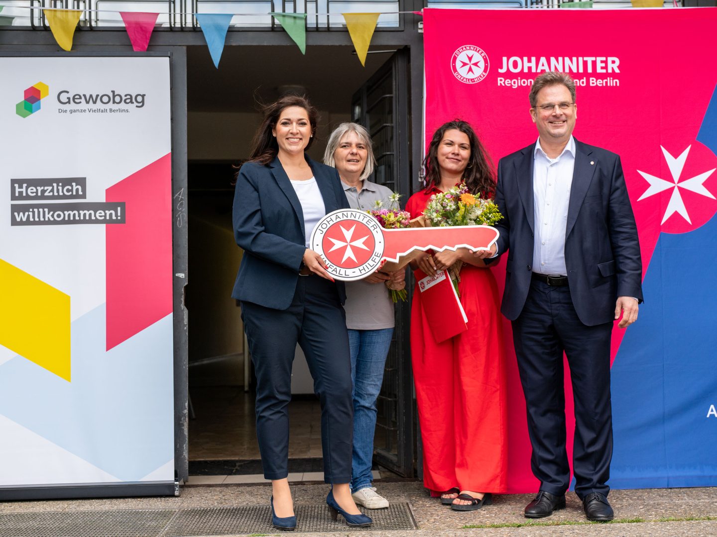 Vier Erwachsene stehen vor bunten Bannern, zwei halten einen übergroßen symbolischen Schlüssel und Blumen – feierliche Eröffnung eines Projekts.