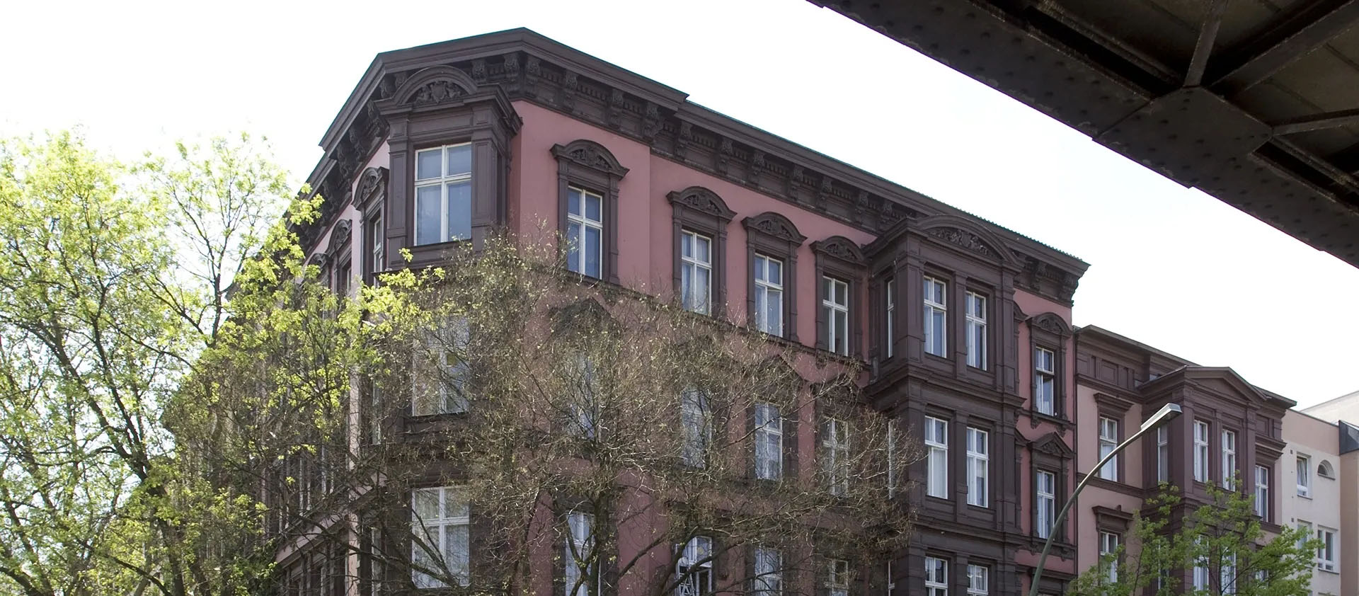 Historisches Wohnhaus mit reich verzierten Fassaden in Rosa und Braun, teils von Bäumen verdeckt, oben angeschnittener Brückenträger.