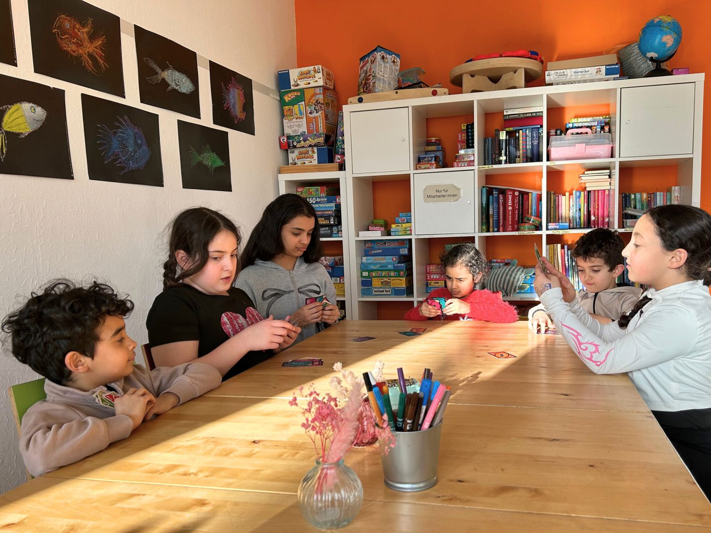 Sechs Kinder sitzen an einem Holztisch in einem bunten Raum und spielen konzentriert Karten, Regale mit Spielen und Büchern im Hintergrund.