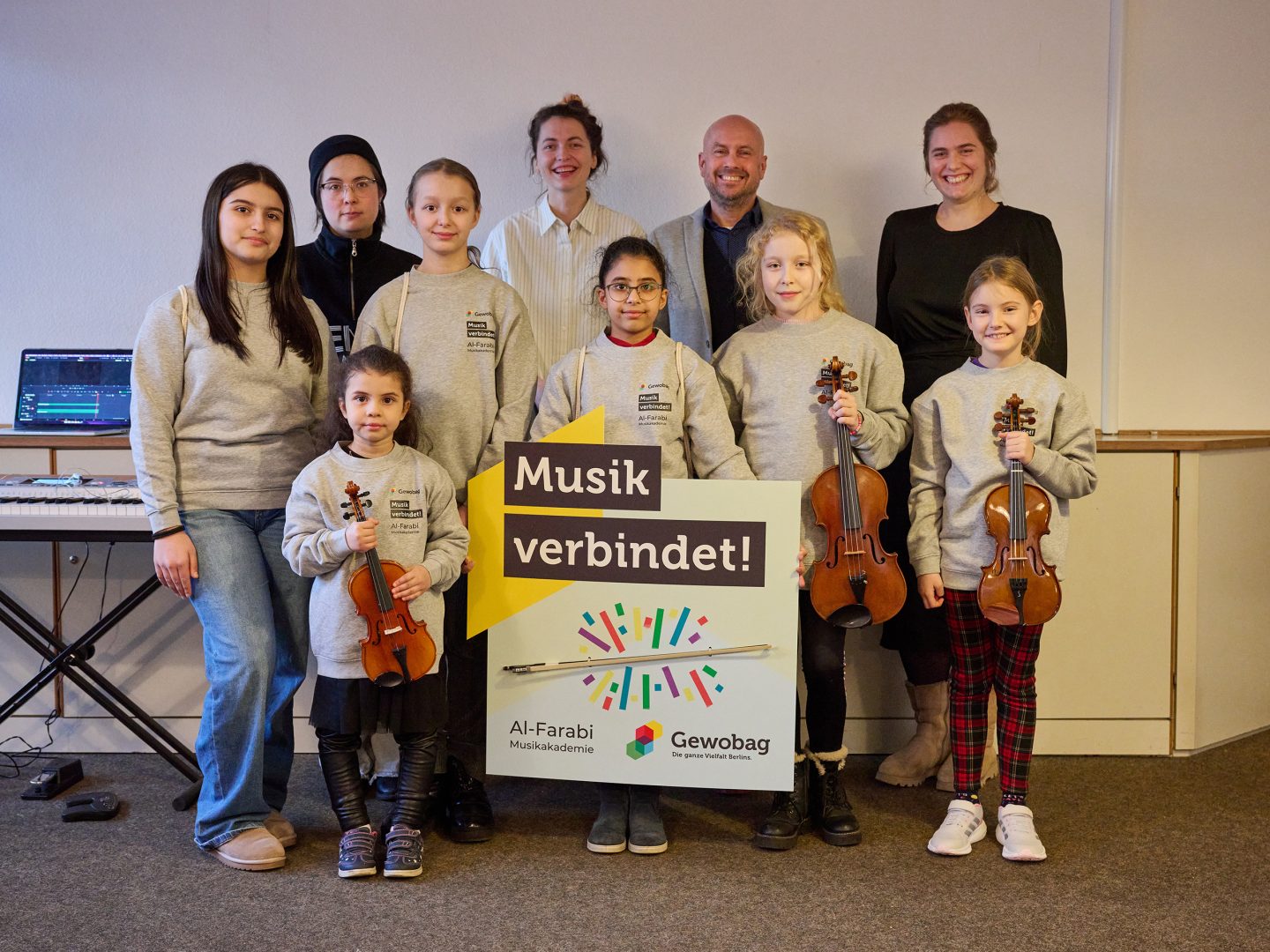 Kinder und Erwachsene der Al-Farabi-Musikakademie posieren im Probenraum mit Violinen hinter einem Schild mit der Aufschrift „Musik verbindet!“.