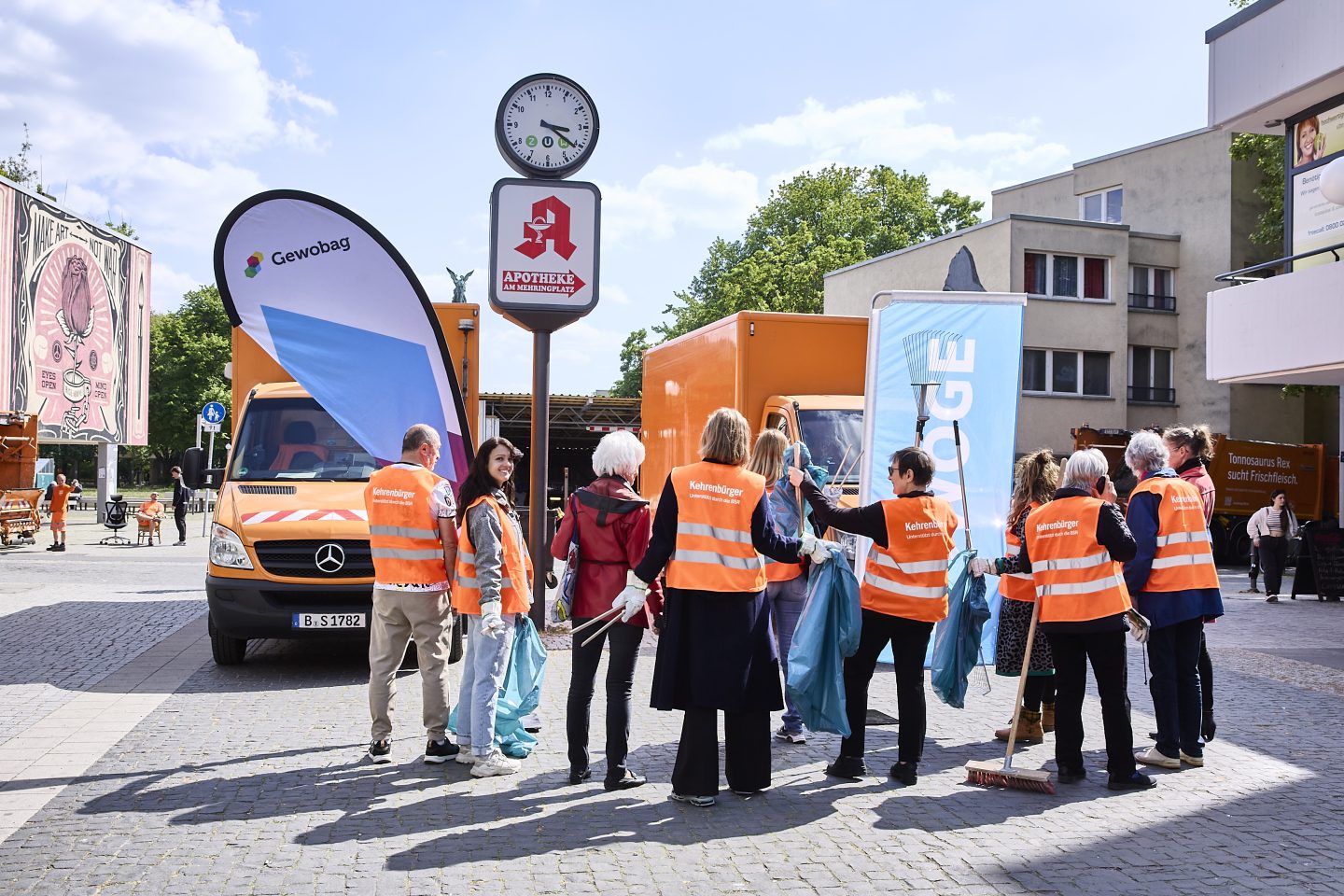 Gruppe Menschen in orange Warnwesten sammelt Müll mit Greifzangen und Säcken vor orangefarbenen Fahrzeugen auf einem Platz mit Uhr und Schildern.