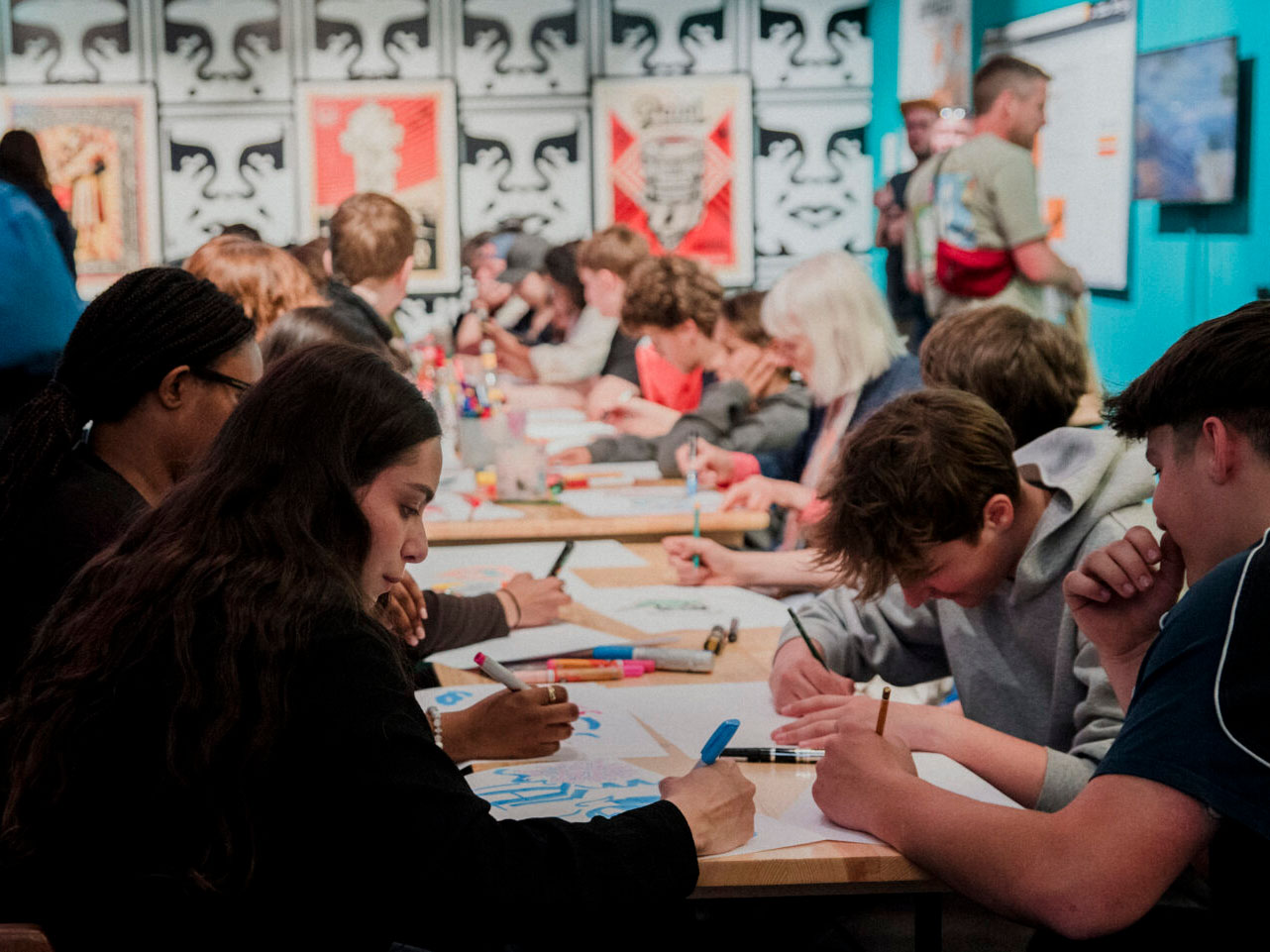 Jugendliche sitzen dicht an dicht an einem langen Tisch und zeichnen konzentriert mit Stiften bei einem kreativen Workshop in Innenräumen.