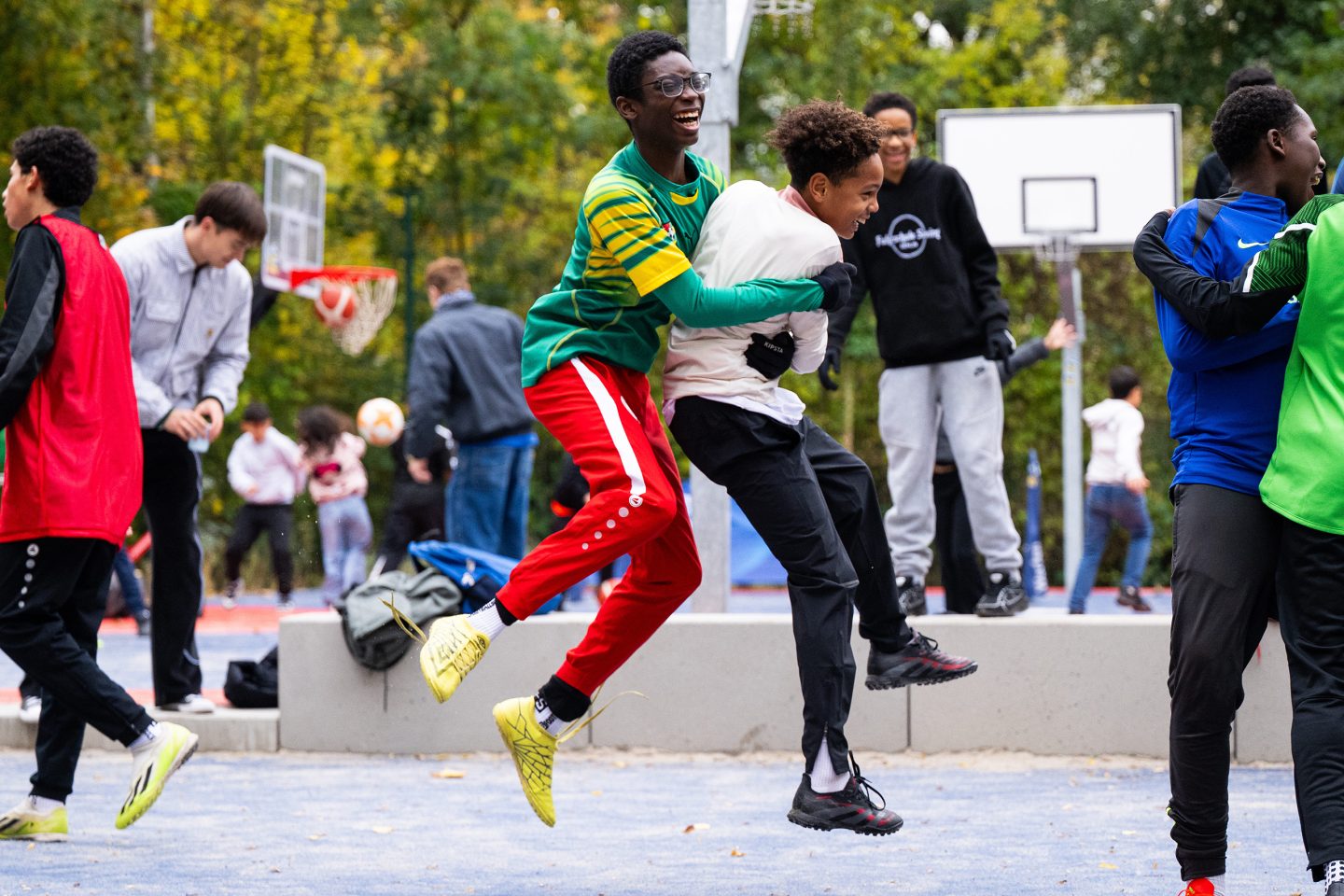 Jugendliche toben lachend auf einem Sportplatz, zwei Jungen springen sich beim Fußballspiel in die Arme, andere spielen im Hintergrund.