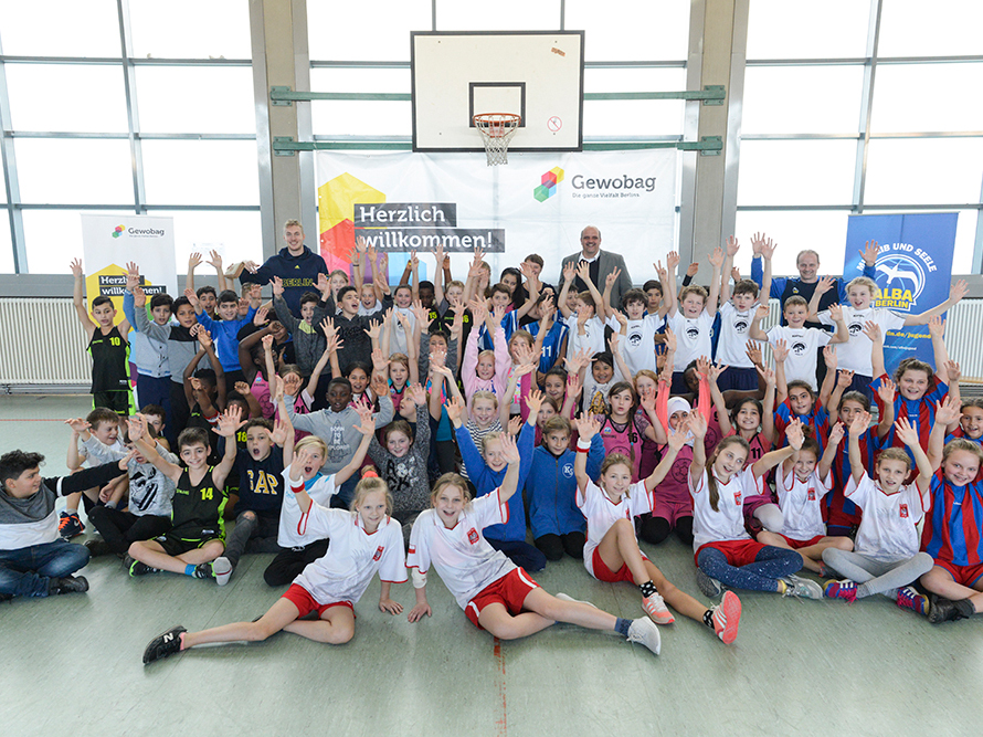 Große Kindergruppe in Sporthalle posiert fröhlich in Sporttrikots, alle heben die Arme; im Hintergrund Banner „Herzlich willkommen!“.