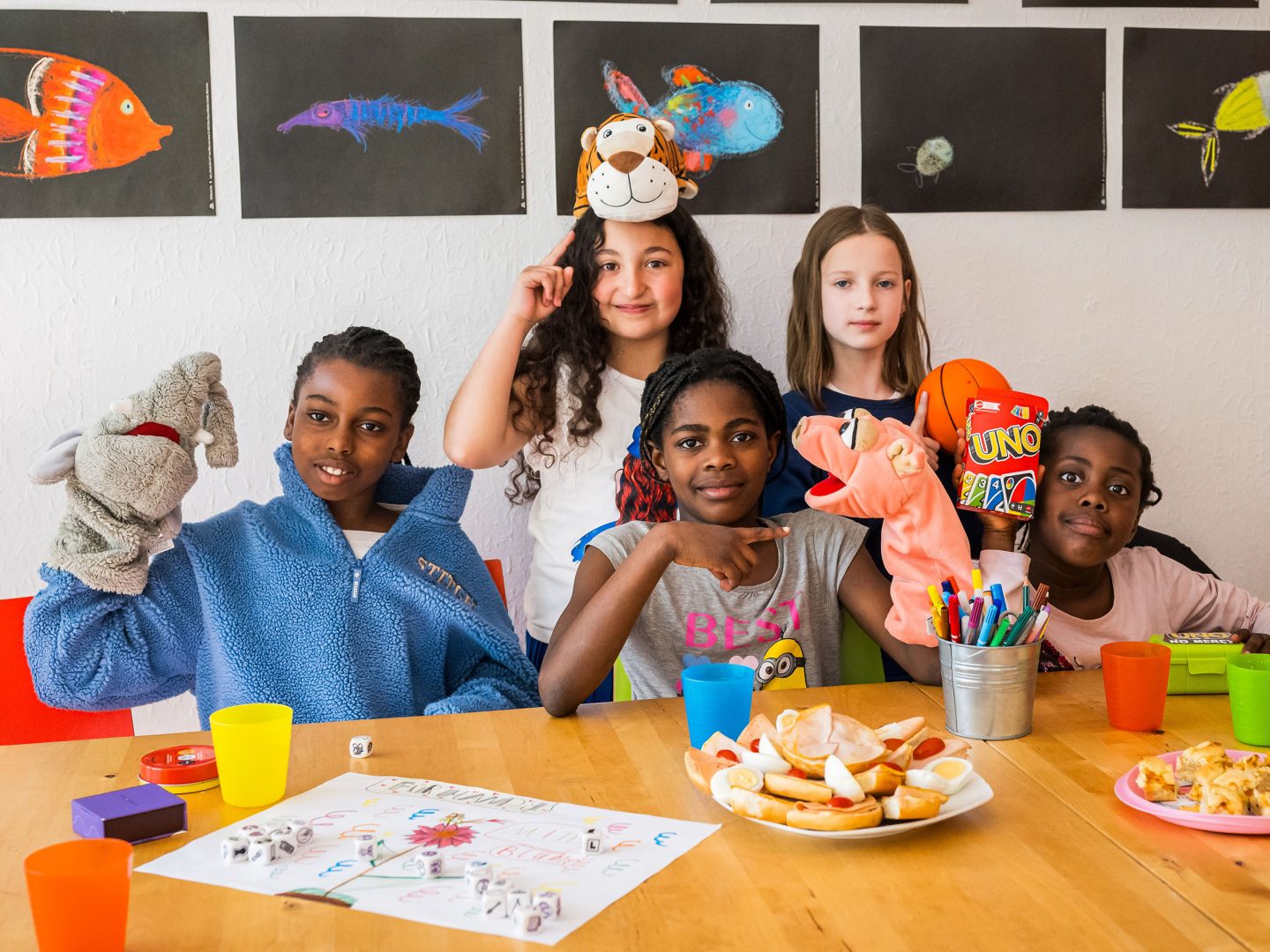 Fünf Kinder sitzen fröhlich an einem gedeckten Tisch, halten Handpuppen und Spiele hoch; bunte Fischzeichnungen hängen an der Wand.