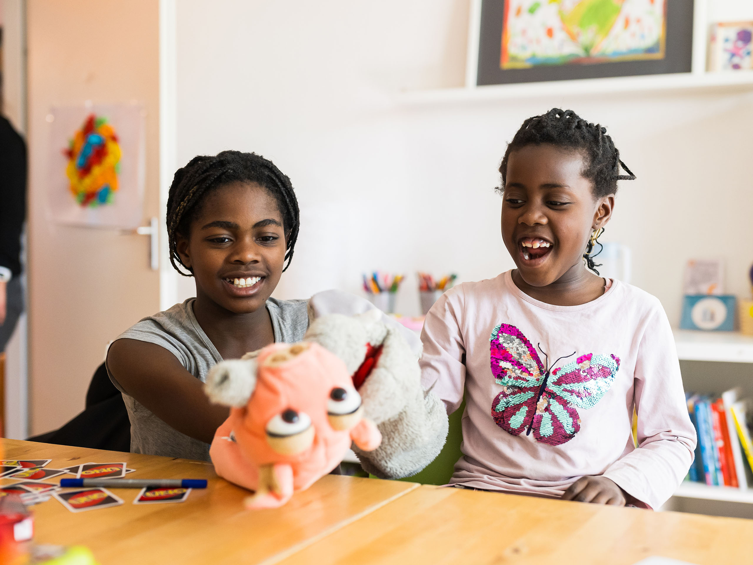 Zwei Mädchen sitzen lachend an einem Tisch und spielen mit bunten Handpuppen; im Hintergrund Kinderzeichnungen und Stifte.