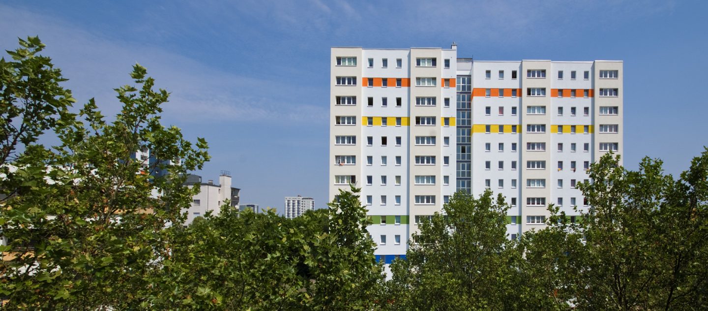 Hohes weißes Wohnhaus mit farbigen Streifen in Orange, Gelb und Grün, umgeben von dichten Baumkronen unter blauem Himmel.