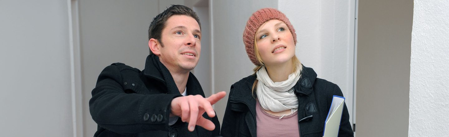 Zwei junge Menschen stehen in einem hellen Flur; der Mann zeigt nach vorne, die Frau mit Mütze und Ordner blickt interessiert nach oben.