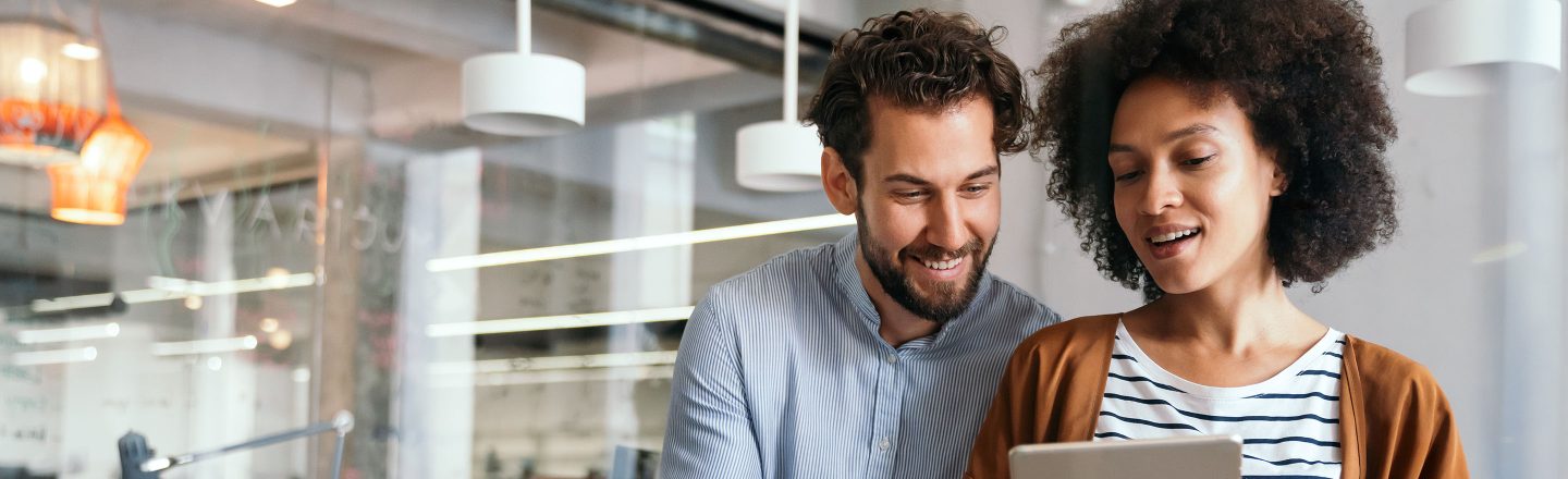 Zwei KollegInnen stehen im modernen Büro und schauen lächelnd gemeinsam auf ein Tablet, das die vordere Person in den Händen hält.