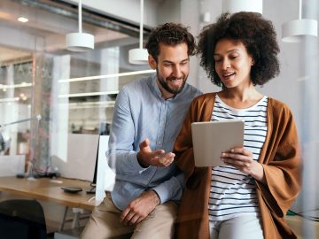 Zwei KollegInnen stehen im modernen Büro und schauen lächelnd gemeinsam auf ein Tablet, das die vordere Person in den Händen hält.