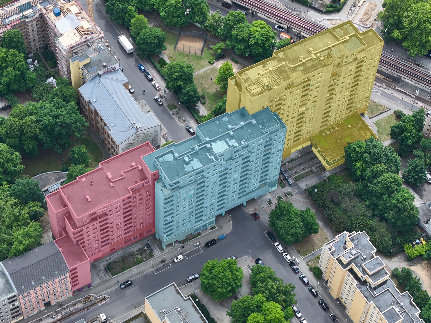 Luftaufnahme eines Stadtviertels mit drei farbig markierten Wohnblöcken: rot, blau und gelb hervorgehobene Hochhäuser zwischen Bäumen.