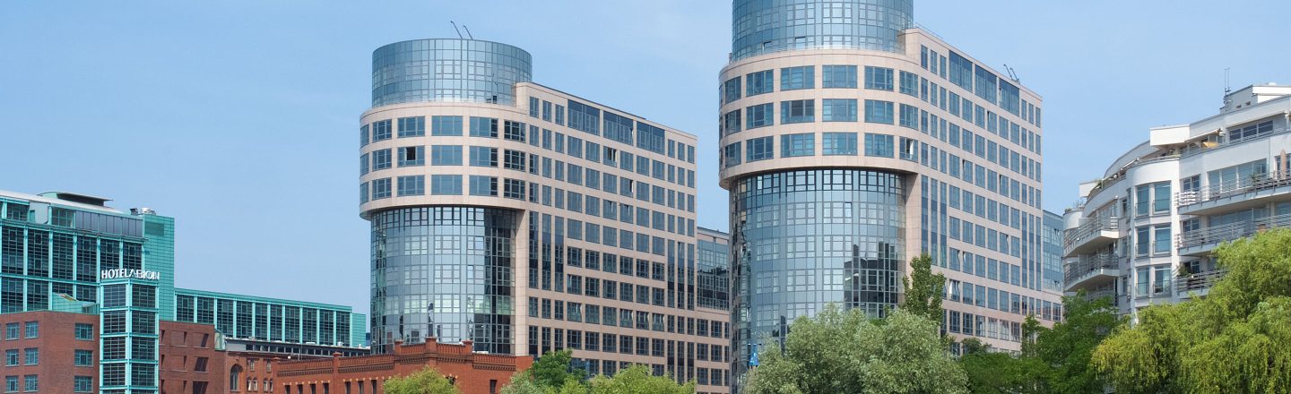 Moderne mehrstöckige Bürogebäude mit großen Glasfassaden vor blauem Himmel, im Vordergrund grüne Bäume.