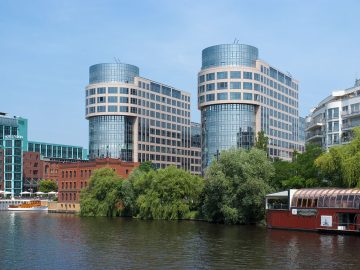 Moderne Bürogebäude mit Glasfassaden am Flussufer, davor dichtes Grün und ein Anleger mit verglastem Restaurantsschiff auf dem Wasser.