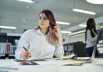 Person schreibt in Büroraum Notizen und blickt nachdenklich zur Seite; vor ihr liegen Bücher und ein aufgeklappter Laptop.