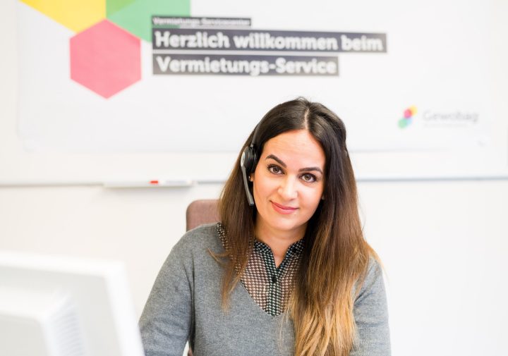 Der Vermietungs-Service für Mietinteressenten Junge Frau mit Headset sitzt an einem Schreibtisch und schaut freundlich in die Kamera; hinter ihr Schild „Herzlich willkommen beim Vermietungs-Service“.