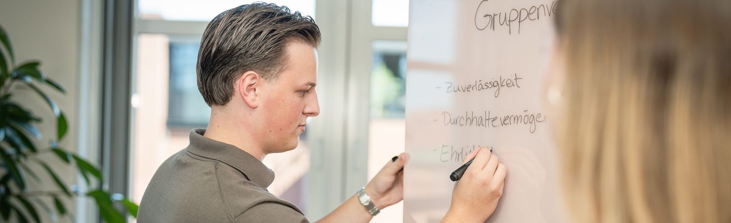 Junger Mann schreibt mit Stift Begriffe wie Zuverlässigkeit und Ehrlichkeit auf ein Whiteboard, eine junge Frau steht daneben.