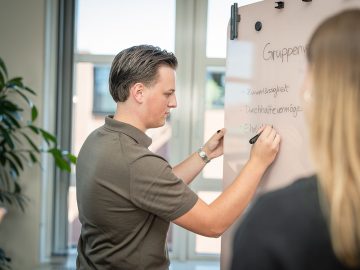 Junger Mann schreibt mit Stift Begriffe wie Zuverlässigkeit und Ehrlichkeit auf ein Whiteboard, eine junge Frau steht daneben.