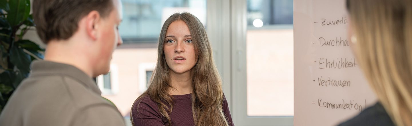 Junge Frau erläutert am Whiteboard Begriffe wie Zuverlässigkeit und Vertrauen, zwei junge Menschen hören ihr aufmerksam im Gespräch zu.