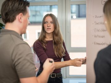 Junge Frau erläutert am Whiteboard Begriffe wie Zuverlässigkeit und Vertrauen, zwei junge Menschen hören ihr aufmerksam im Gespräch zu.