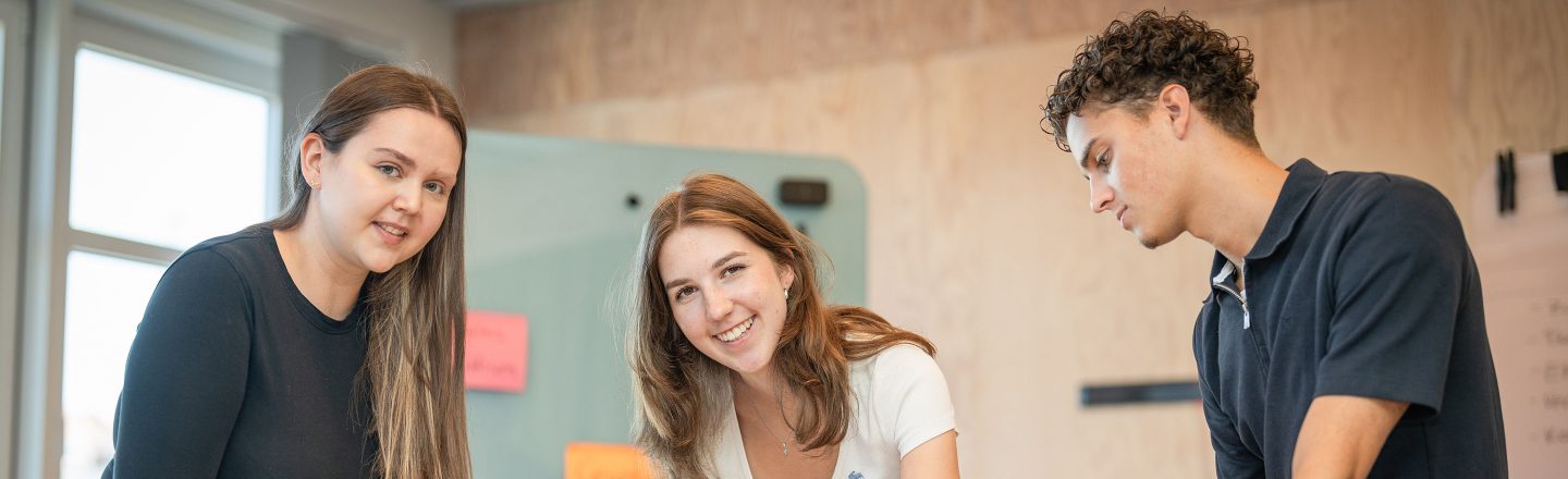 Drei junge Menschen arbeiten zusammen an einem Tisch im hellen Raum; zwei lächeln in die Kamera, eine Person konzentriert nach unten.