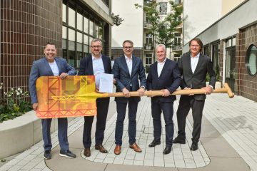 Fünf Männer in Anzügen stehen in einem Innenhof und halten gemeinsam einen großen, bunt gestalteten symbolischen Schlüssel.